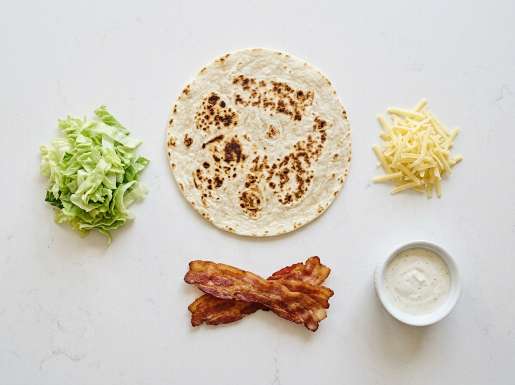Overhead flat lay of Cook Out Bacon Wrap ingredients: flour tortilla, crispy bacon strips, shredded cheddar cheese, shredded lettuce, and ranch dressing in a small bowl