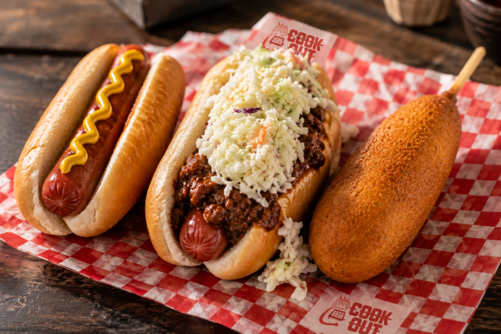 Three Cook Out hot dog varieties side by side — plain hot dog, chili slaw dog with beef chili and coleslaw, and golden corn dog on a stick